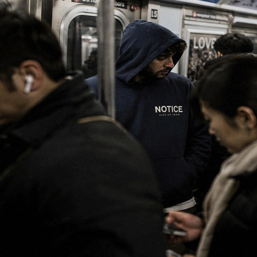 S-001 Closed Eye hoodie front with NOTICE and Kids Of Iran chest print, worn on a subway train